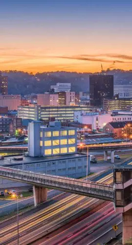 A vibrant cityscape is captured at dusk, featuring illuminated buildings and intersecting highways against a backdrop of rolling hills and a colorful sky.
