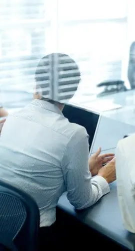 Two professionals are engaged in work at their computers in a modern office setting, with a view through a glass partition.