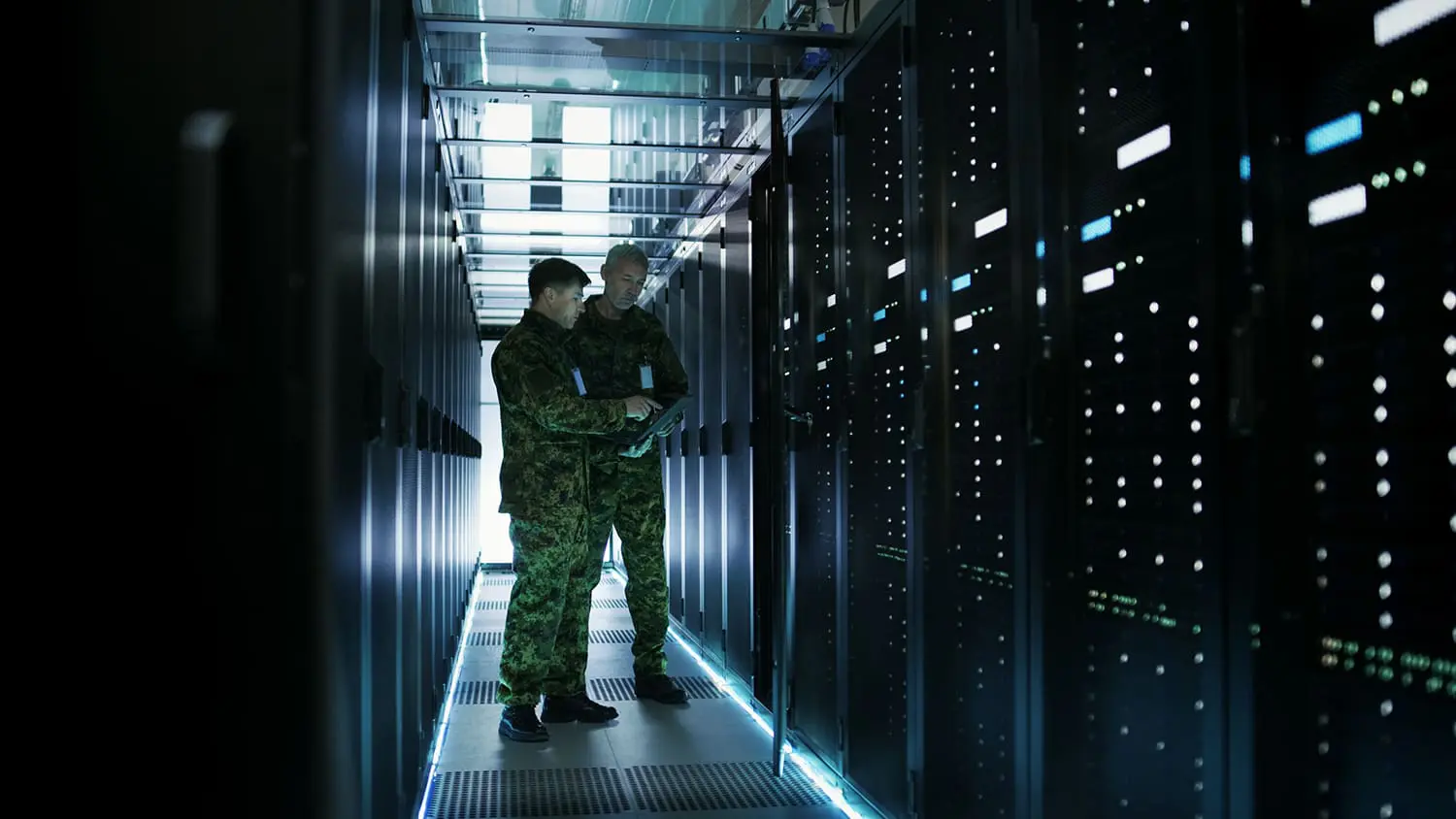 A person in military camouflage stands in a data center corridor lined with server racks illuminated by blue lights.