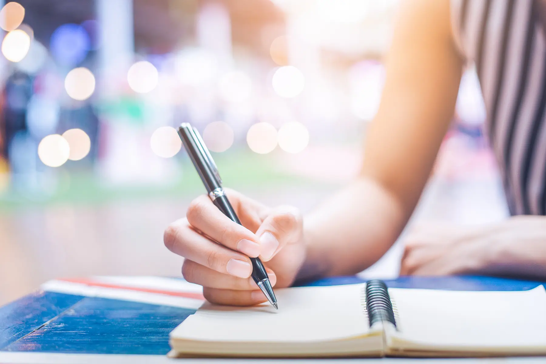 A person is writing in a notebook on a desk with a blurry background that suggests a bokeh effect typically associated with lights.