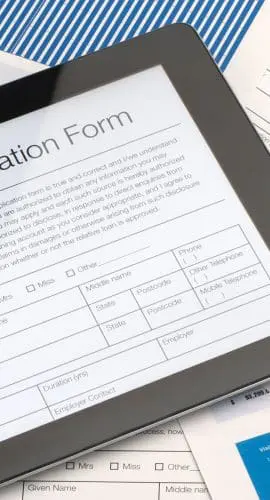 A tablet displaying a mortgage application form is placed on a desk cluttered with documents, a pen, eyeglasses, and a black notebook.