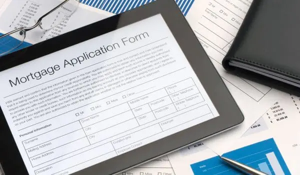 A tablet displaying a mortgage application form is placed on a desk cluttered with documents, a pen, eyeglasses, and a black notebook.