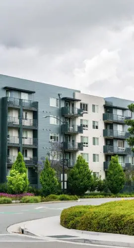 A modern mid-rise apartment building is surrounded by greenery and trees, with a curved road in the foreground.