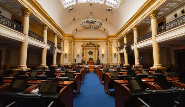 A legislative assembly hall with empty desks and a large chandelier overhead.