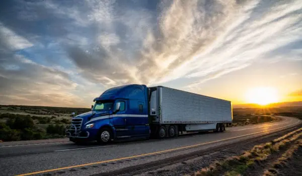 A blue semi-truck drives on a highway at sunset with a dramatic sky in the background.