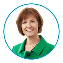 A woman with short brown hair is wearing a green jacket and a pearl necklace, framed by a blue circle against a white background.