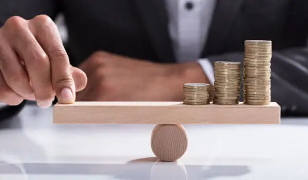 Close-up Of A Businessperson's Hand Balancing Stacked Coins On Wooden Seesaw With Finger Over Desk