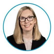 A person with blonde hair, wearing glasses and a black blazer with a light-colored top, is smiling inside a blue circular frame.