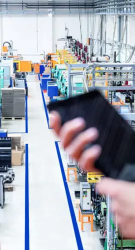 Horizontal color image of businesswoman - unrecognizable person - working with digital tablet in large futuristic factory. Woman standing on top of a balcony, holding touchpad and checking inventory of a manufacturing company on touchscreen tablet. Focus on futuristic machines in background, businesswoman's hands holding black tablet defocused.