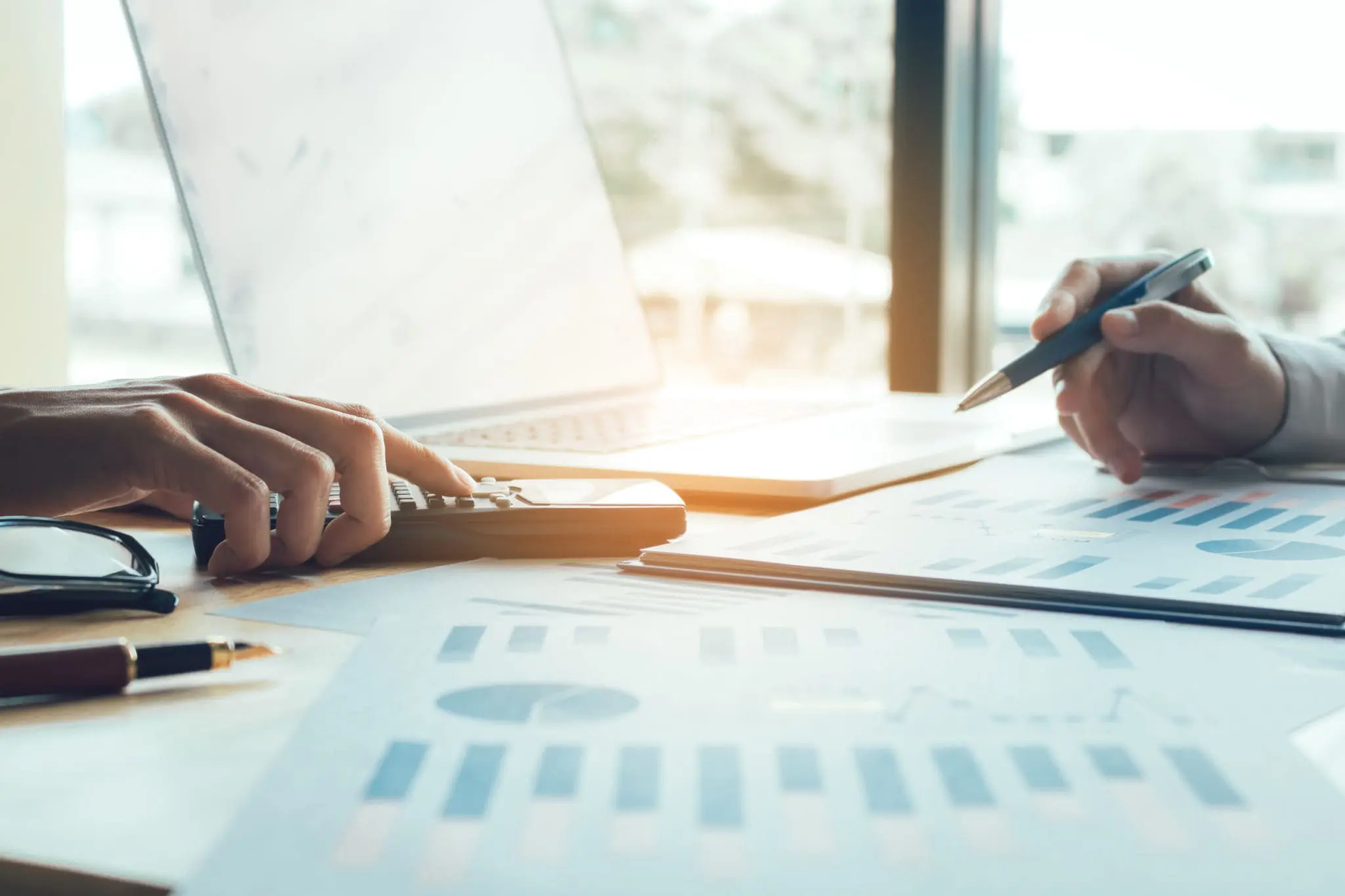 Two people are working on financial documents and using a calculator at a desk with a laptop in the background.
