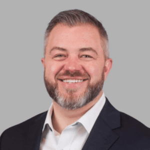 A smiling person with a beard is wearing a dark blazer and a light-colored shirt against a plain background.