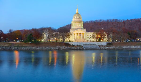 A grand, illuminated capitol building at twilight reflects elegantly on the calm waters of a nearby river.
