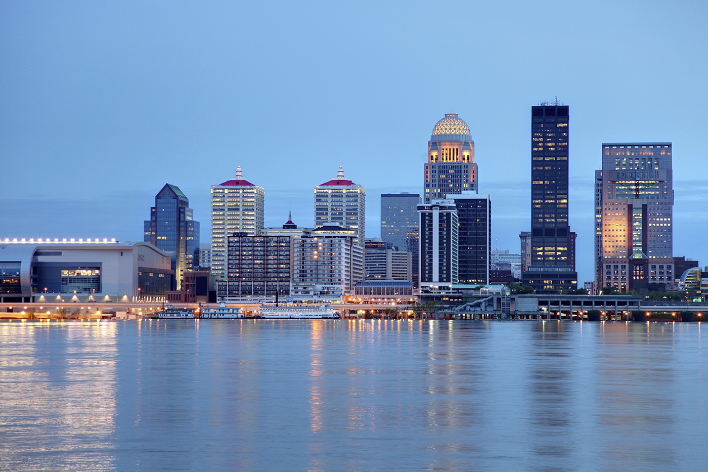 A skyline of a modern city with illuminated buildings reflected in a large body of water during twilight.