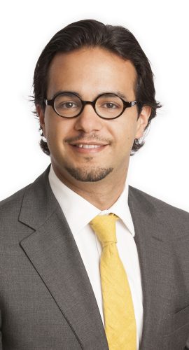 A professional-looking individual with glasses is wearing a gray suit, a white shirt, and a yellow tie, and is smiling at the camera.