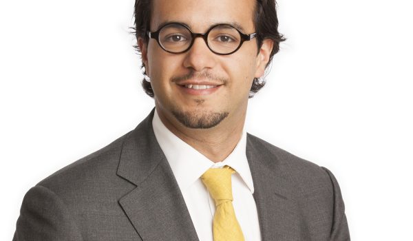 A professional-looking individual with glasses is wearing a gray suit, a white shirt, and a yellow tie, and is smiling at the camera.