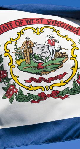 A close-up of the West Virginia state flag featuring its seal with a red ribbon and its motto, amidst folds of wavy fabric.