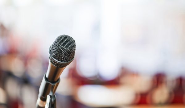 A microphone is in focus against a softly blurred background, suggesting a public speaking or performance setting.