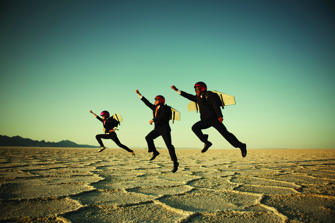 Individuals are seen joyfully leaping across a cracked desert surface under a vast sky at what appears to be either sunrise or sunset.