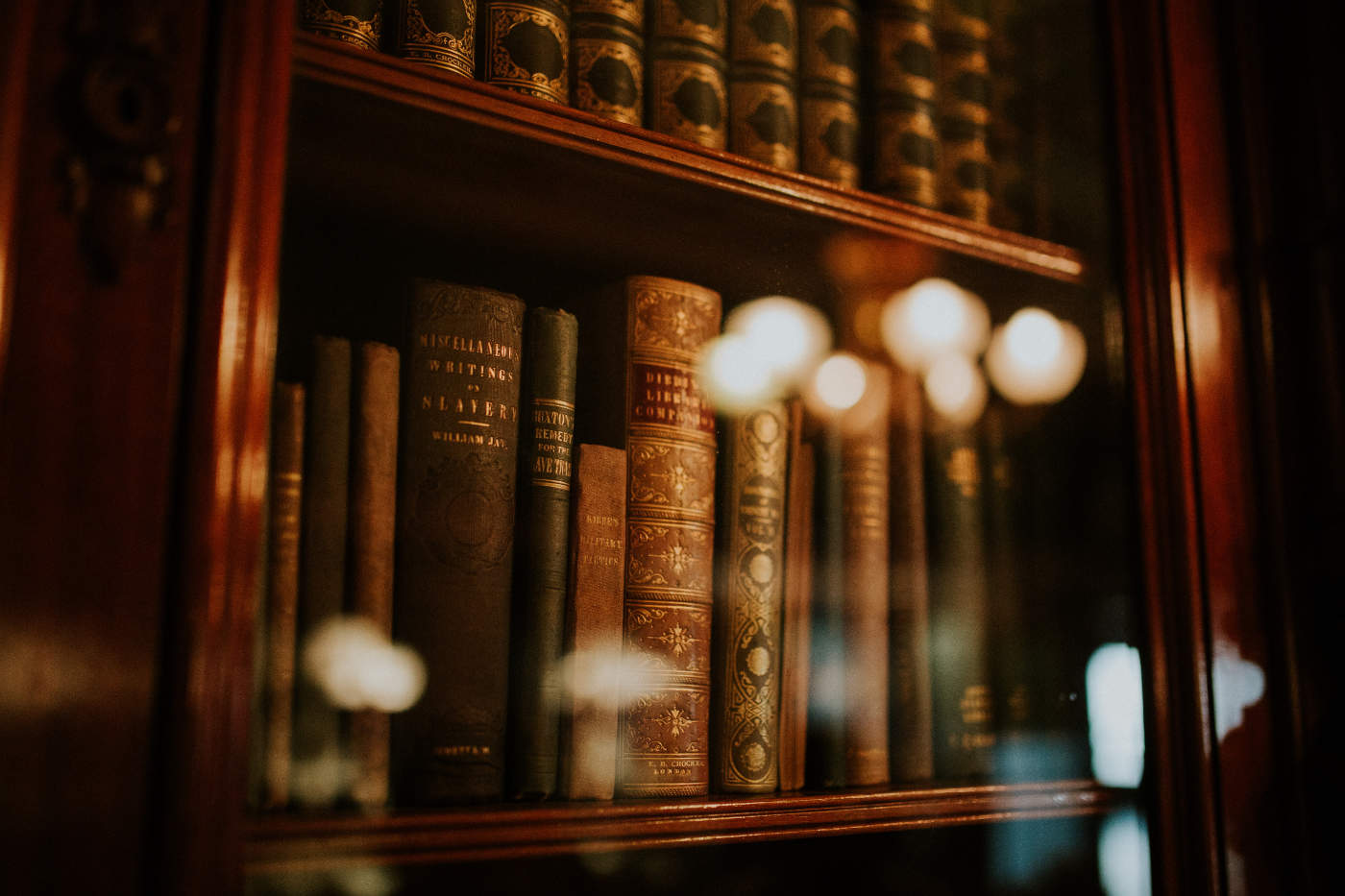 A collection of old books with gold lettering on the spine is displayed in a wooden bookcase behind glass, with soft, warm lights reflecting off the surface.