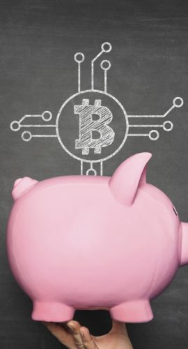 A pink piggy bank is being held up against a chalkboard background with a Bitcoin and circuit diagram drawn above it.