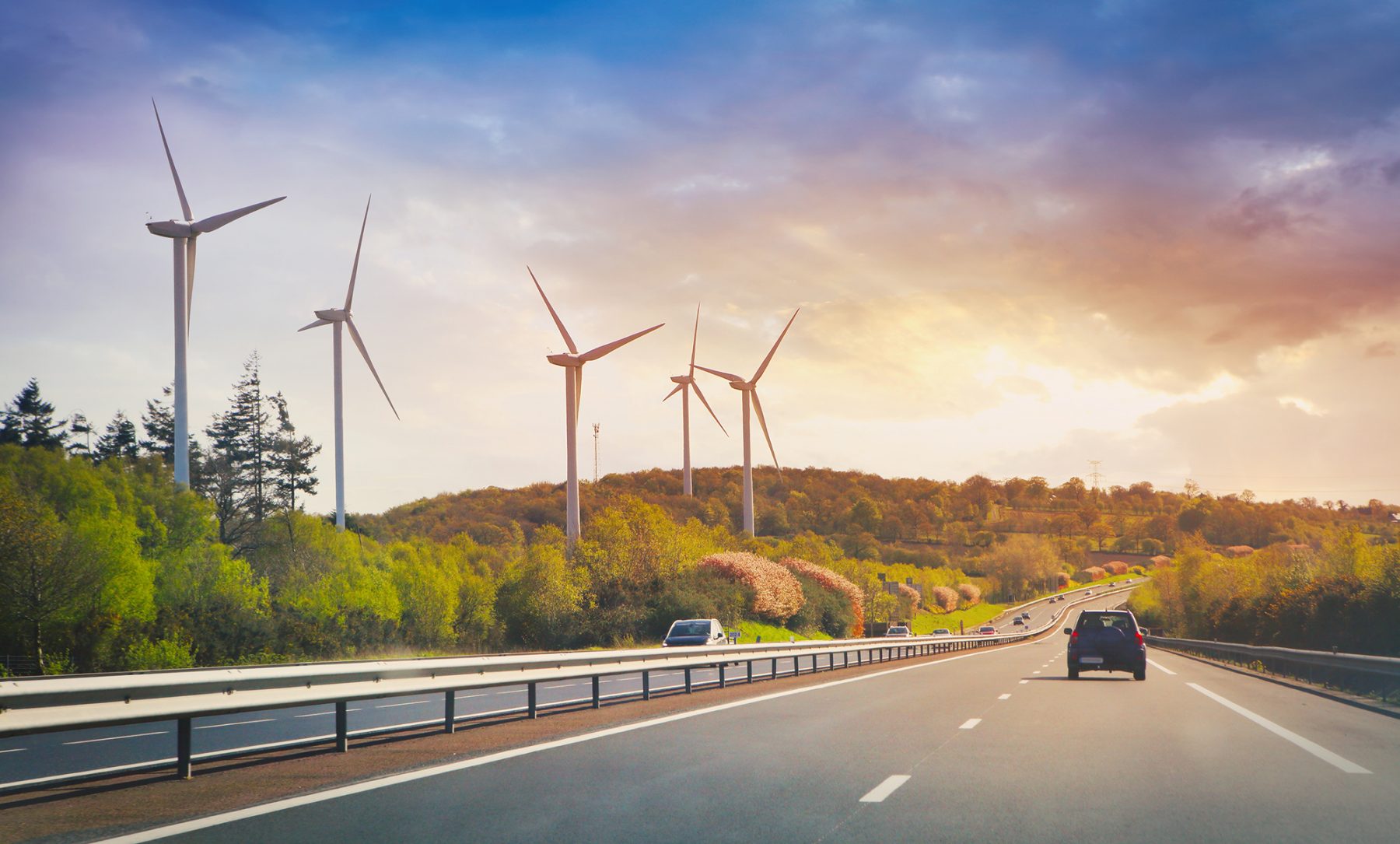 A highway leading towards wind turbines against a backdrop of a colorful sky and lush greenery.