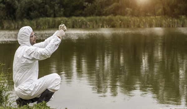 A person in a protective suit is kneeling by a body of water, possibly collecting a sample or examining the environment.