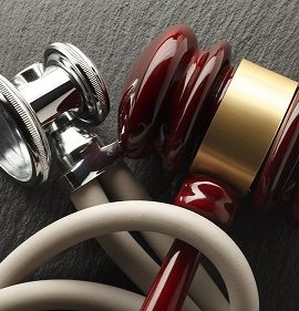 A red and a gold stethoscope with white tubing are resting on a textured surface.