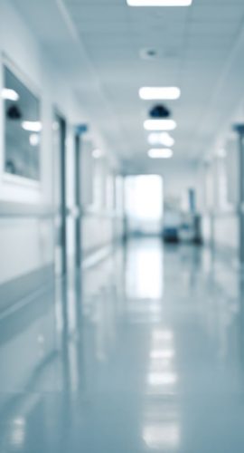 A blurry figure is visible at the end of a bright, clean hospital corridor with white walls and a reflective floor.