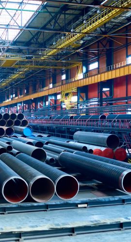 A collection of large industrial pipes is stored in a spacious factory with a high ceiling and overhead cranes.