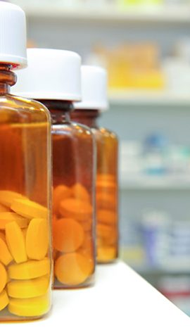 Orange prescription bottles filled with pills are lined up on a pharmacy shelf, with a focus on the closest bottle and a blurred background.