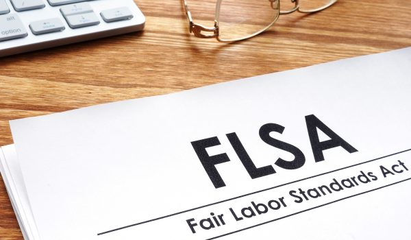 A document titled "FLSA: Fair Labor Standards Act" is placed on a wooden desk next to a pair of glasses and calculator keys.