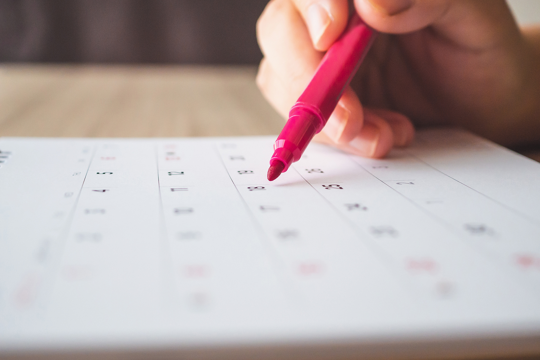 A hand is using a red pen to mark a date on a calendar.