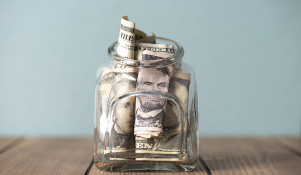 A glass jar filled with rolled and folded U.S. dollar bills sits on a wooden surface against a plain, light-colored background.