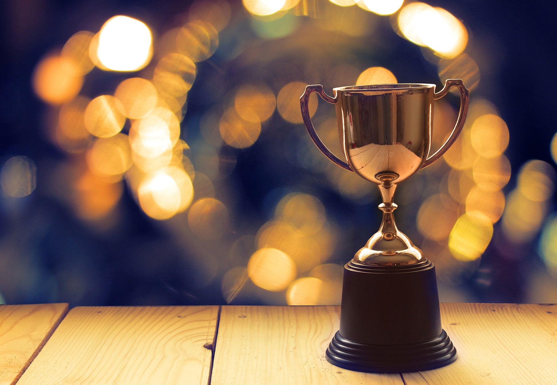 A gold trophy is displayed on a wooden surface with a blurred, bokeh-lit background.