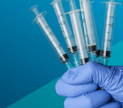 A gloved hand is holding several empty syringes against a blue background.