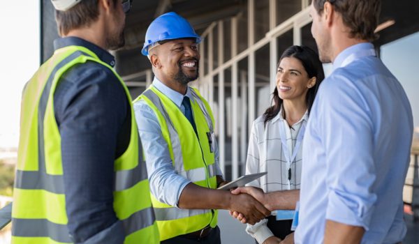 Smiling engineer shaking hands at construction site with happy architect. Handshake between cheerful african construction manager with businessman at bulding site. Team of workers with architects and contractor conclude an agreement with safety uniform.