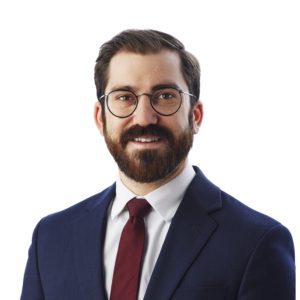 A man with glasses, a beard, and a maroon tie is posing in a business suit against a white background.