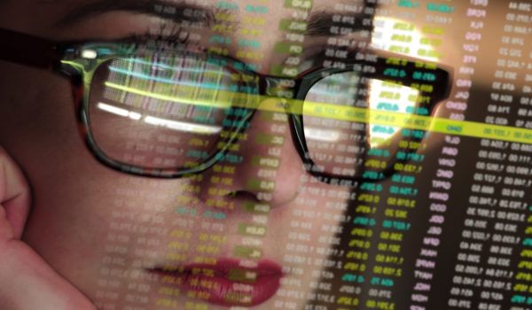 Stock photo of a beautiful young woman’s face very close up. She’s looking at a complicated set of holographic, see-thru numbers. The numbers are also reflected in her spectacles.