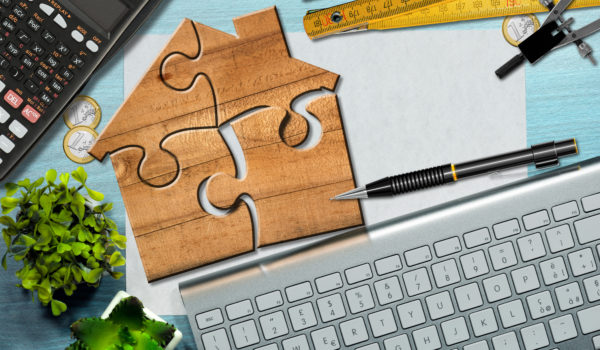 Wooden jigsaw puzzle pieces forming a house shape, on a desk with blank sheet of paper with copy space, calculator, folding ruler, drawing compass, Euro coins, pencil and a computer keyboard.