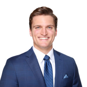 A person in a blue suit and tie with a white shirt smiles against a white background.