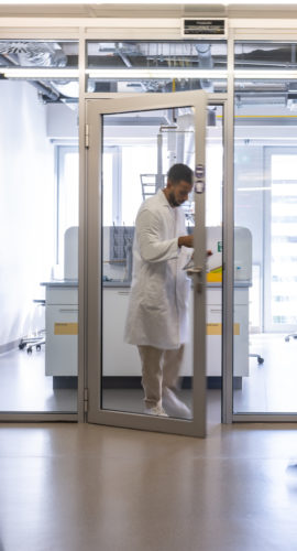 A chemical worker in a white coat stands between the doors of the faculty laboratory, students rush past him in the corridor. Blurred people while moving on the photo.