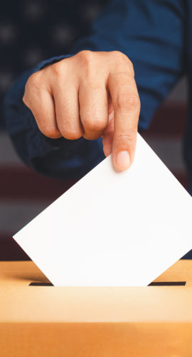 Hand voter holding ballot paper putting into the voting box at place election with an American flag background. Freedom democracy concept. Close-up photo