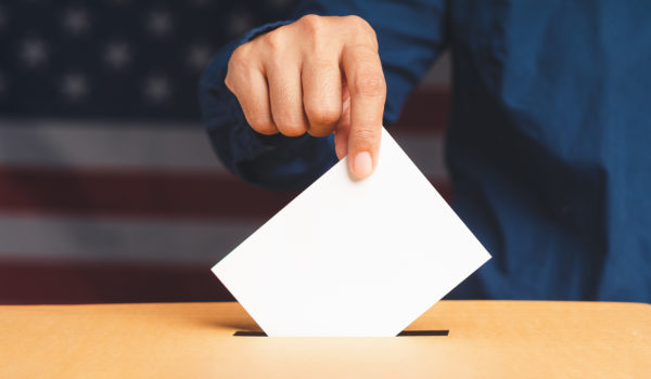 Hand voter holding ballot paper putting into the voting box at place election with an American flag background. Freedom democracy concept. Close-up photo